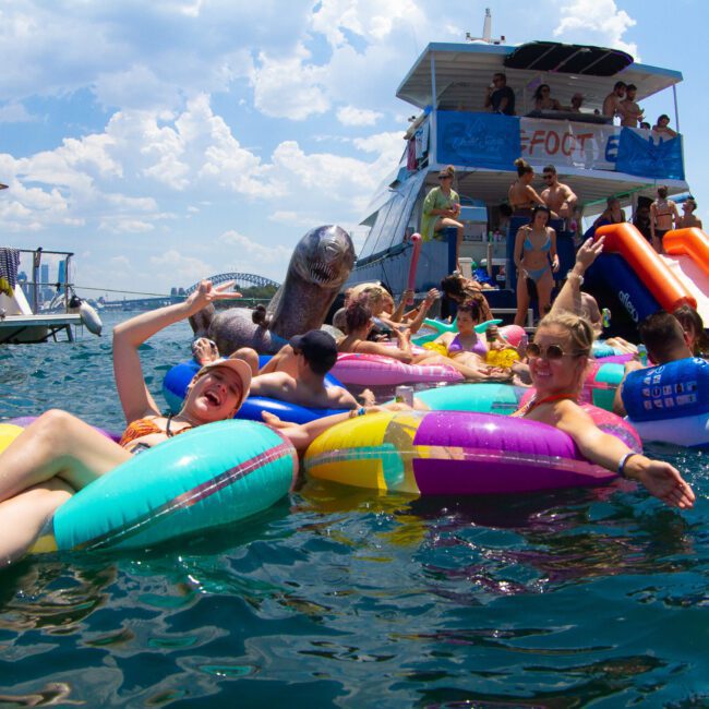 A group of people enjoying a sunny day on an assortment of colorful inflatables in the water, with a large boat in the background. Some people are lounging and smiling, while others are actively playing and sliding off the boat into the water, making for an idyllic summer scene.