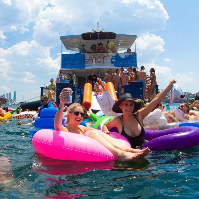 People floating on colorful inflatable tubes in the water, enjoying a sunny day. A large boat with a "foot" sign is in the background, where more people are standing, dancing, and cheering. The blue sky is scattered with clouds, adding to the lively and festive atmosphere.