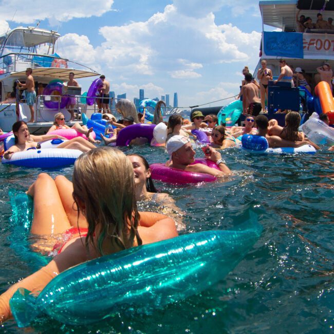 A crowd of people enjoys a summer day in the water, surrounded by colorful inflatables and floating near two anchored boats. The sky is partly cloudy, and several people onboard are preparing to dive into the refreshing water.