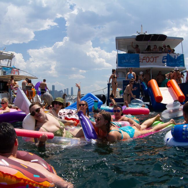 A lively group of people float in the water on various inflatable rafts, including a donut and a unicorn. Two boats are anchored nearby, with people socializing onboard. The sky is partly cloudy, and the atmosphere remains festive and joyful as everyone enjoys the warm day on the river.