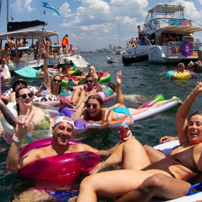 A lively group of people enjoys a sunny day on the water. They float on various inflatables and inner tubes, with two boats anchored nearby. The skyline of a scenic city can be seen in the background, and everyone appears to be smiling and having a great time.