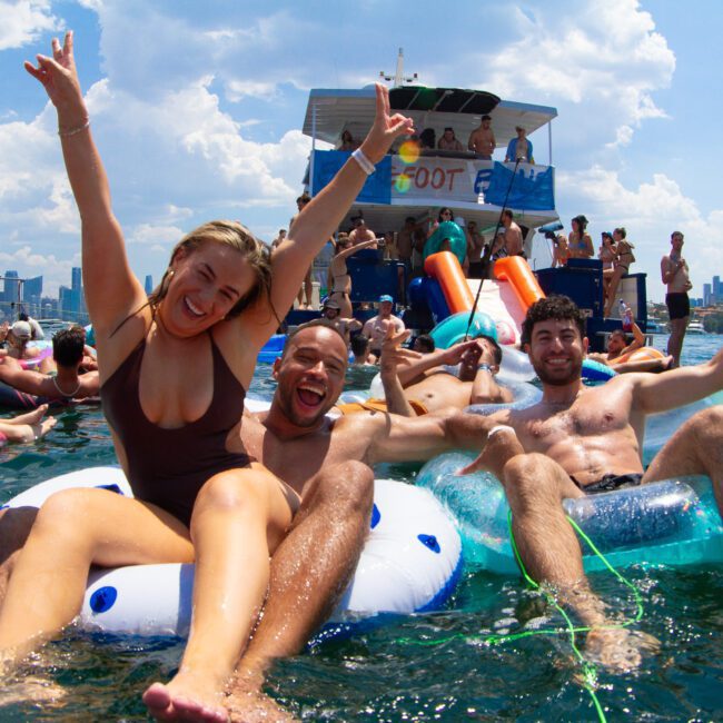 A group of people enjoying a sunny day on the water. Three friends in the foreground are sitting on inflatable tubes, smiling, with one person raising their arms in celebration. In the background, more people are relaxing on inflatable floats near a double-deck boat.