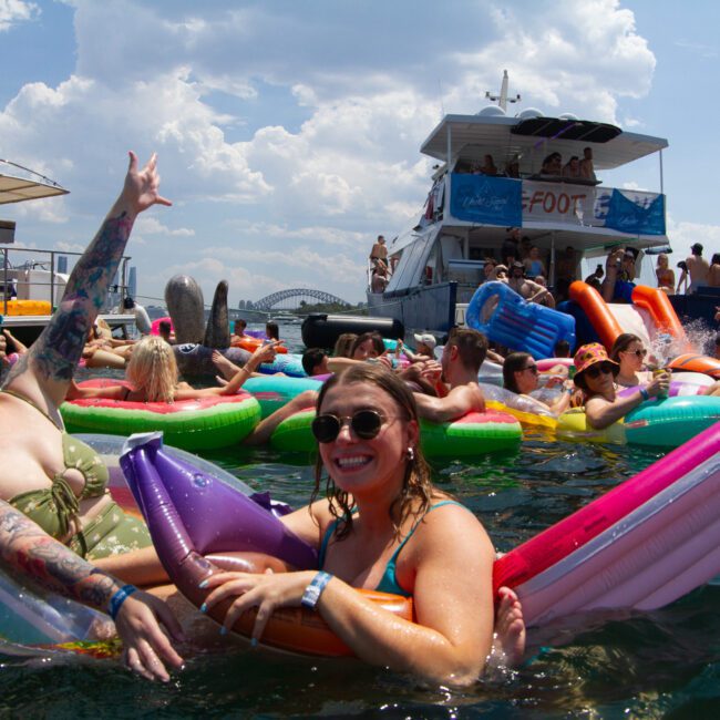 People are savoring a sunny day on the water, floating on vibrant inflatable rafts near two large boats. Several individuals are smiling and waving, with upbeat music playing and others dancing on the boats. The backdrop includes a partly cloudy sky and a scenic distant bridge.