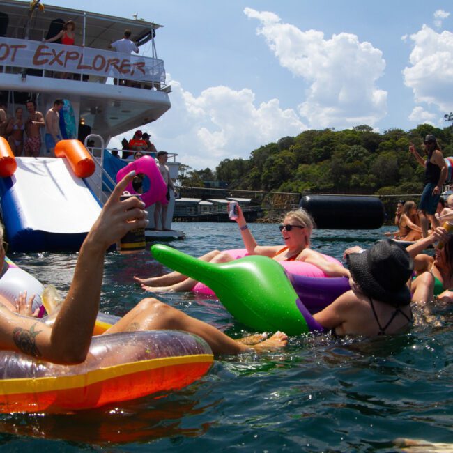 A lively group of people float in an assortment of colorful inflatable rafts in a scenic body of water. In the background, a large boat named "Barefoot Explorer" is docked, with more people enjoying the sunshine and water slides. A stunning mountain range enhances the vibrant atmosphere.
