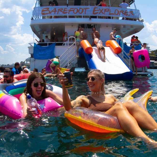 People floating on colorful inflatables in a body of water, partying near a large boat with 'Barefoot Explorer' written on it. Some are in the water, others are on the boat’s slides and enjoying an epic adventure. The weather is sunny with blue skies and scattered clouds.