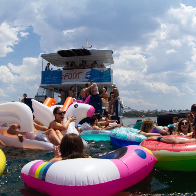 A lively scene of people enjoying a pool float party in the water, with a unicorn and various colorful inflatables, between two anchored boats under a partly cloudy sky. The festive atmosphere is enhanced by the cheerful decorations and laughter echoing across the water.
