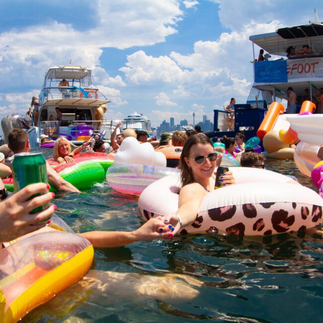 People are enjoying a summer day on a lake, floating on colorful inflatables and holding drinks. They are surrounded by boats under a partly cloudy sky. The atmosphere is vibrant with socializing, relaxation, and excitement as everyone takes in the lively waterfront scene.