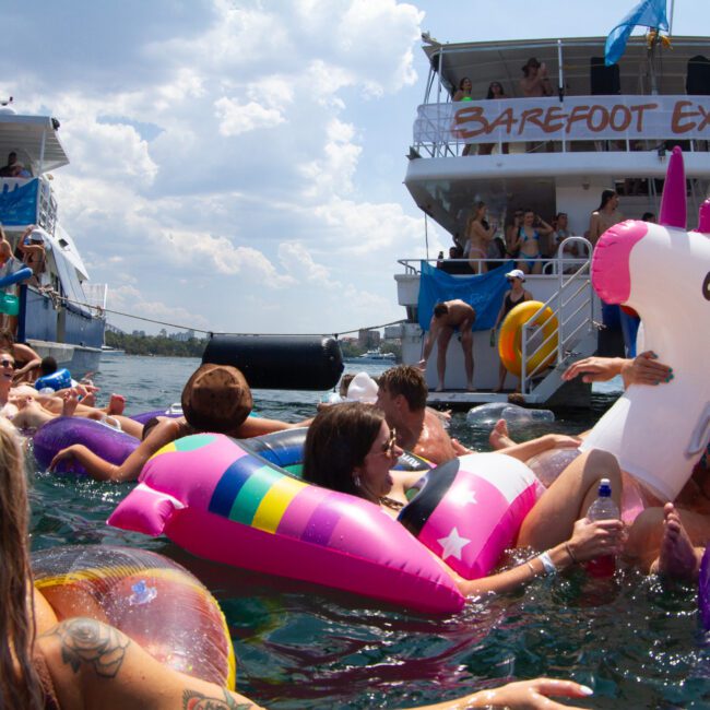 People in swimsuits are having fun in a crowded water area, floating on colorful inflatable toys like unicorns and pools. In the background are two boats with banners that read "Barefoot Explorer" and people observing from decks. The sky is partly cloudy, creating a perfect day for relaxation.
