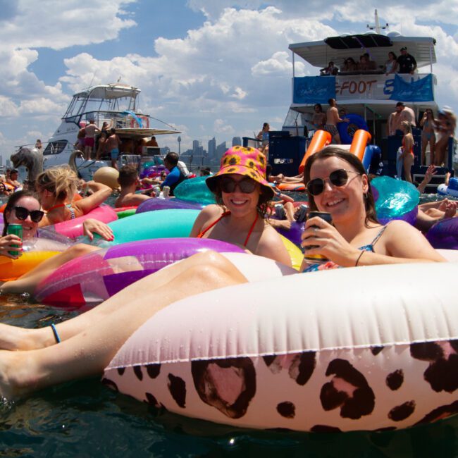 A group of people floating on colorful inflatable tubes in a lake with a party boat in the background on a sunny day. The atmosphere is festive with blue skies, scattered clouds, and water skiers. Some are holding drinks and wearing sunglasses and hats.