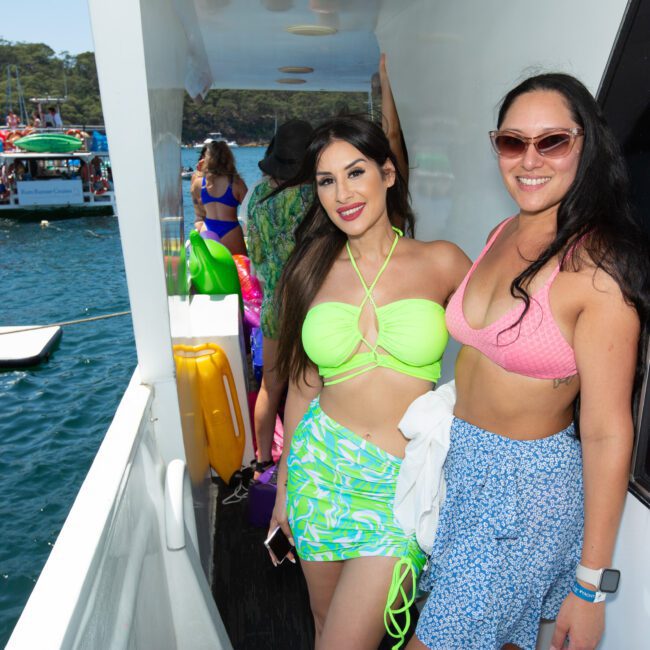 Two women are on a boat, smiling at the camera. One wears a neon green bikini top and patterned skirt, the other a pink bikini top and a blue skirt. It's a bright, sunny day with clear blue water and other boats in the background. They look happy as they cruise through picturesque waters.