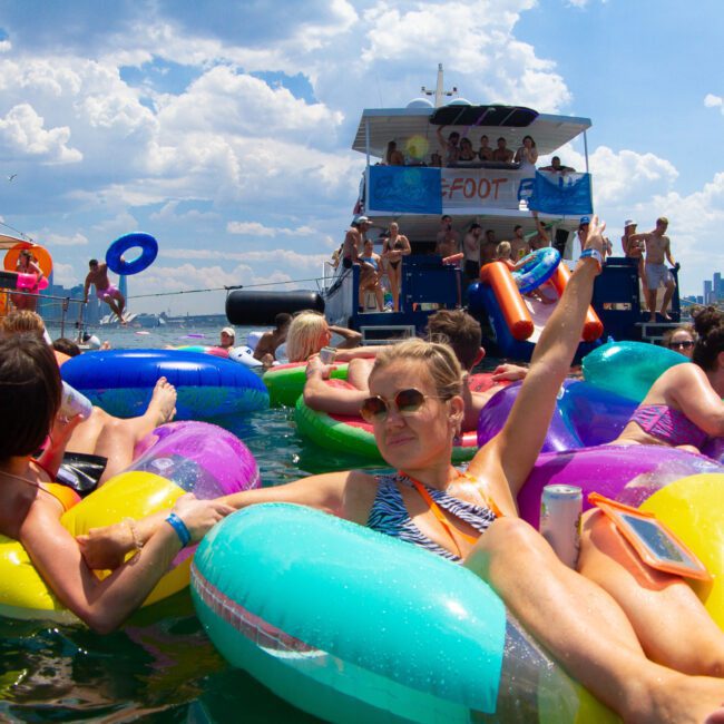 People are enjoying a sunny day on a lake while lounging on colorful inflatable tubes. In the background, there's a double-decker boat with more people socializing and partaking in water sports. The scene is lively and festive, with clear blue skies and fluffy clouds overhead.