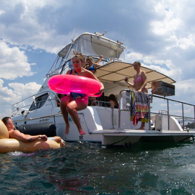 A group of people enjoy a sunny day on a yacht and in the water. One person in a pink swimsuit is mid-air, diving off the yacht while holding a pink inflatable ring. Others are lounging on colorful inflatable floats and standing on the deck of the yacht, savoring their adventure at sea.