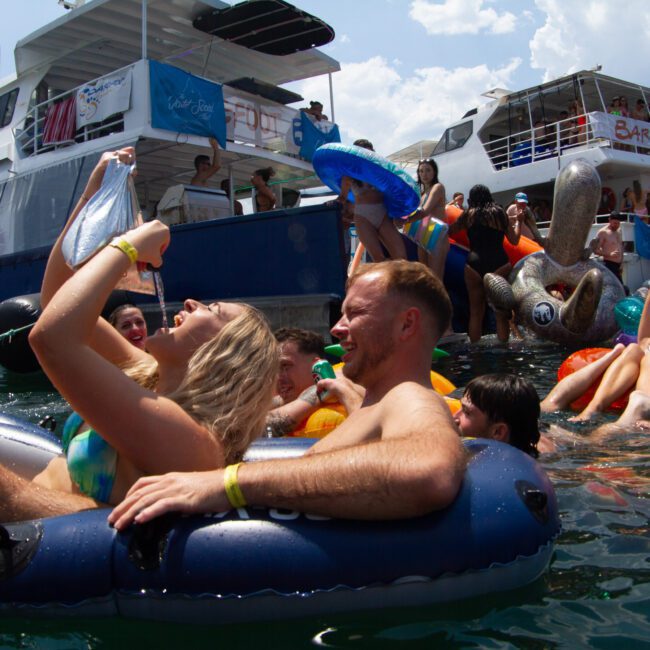 People are enjoying a lively poolside party, surrounded by inflatable tubes and floats. Boats are docked in the background, with banners indicating the event. Participants are socializing, relaxing, and having fun under a sunny sky while a DJ keeps the music pumping.