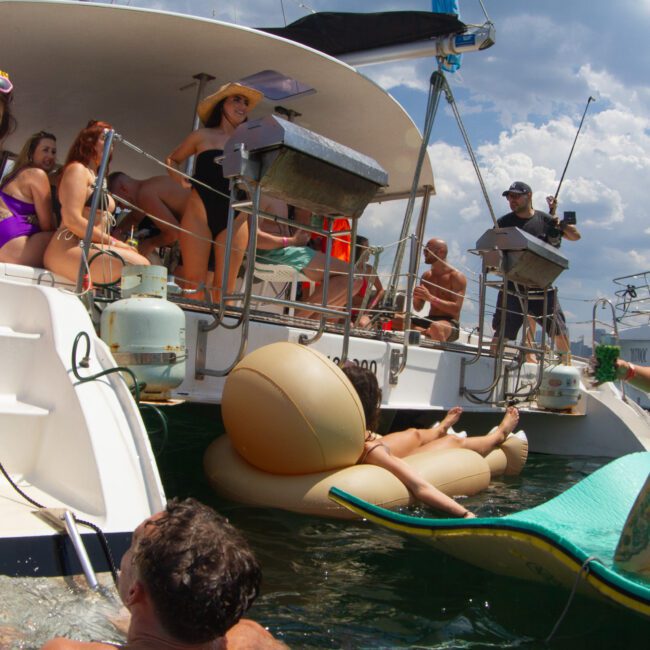 A group of friends are enjoying a sunny day on a boat. Some are on the deck, relaxing and chatting, while others float in the water on inflatable loungers. Everyone is wearing swimsuits, and the atmosphere is lively and fun.