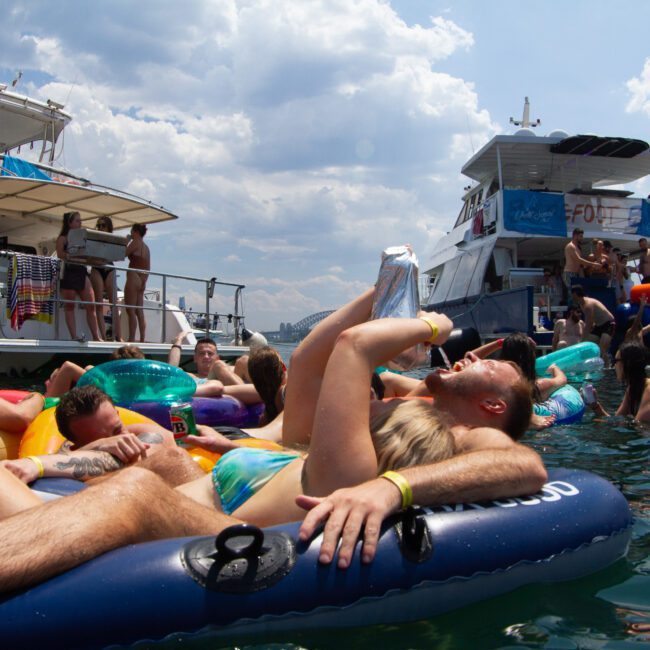 A group of people relaxes on inflatable floats and rafts in the water, surrounded by two anchored boats on a sunny day. Some are drinking and socializing, while others are lounging. The sky is partly cloudy, and in the background, majestic mountains are visible.