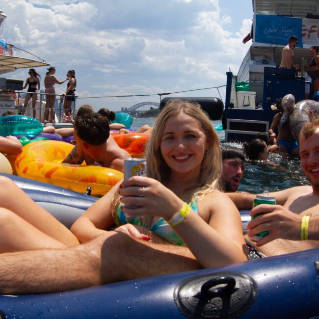 A group of friends enjoying a sunny day on the water, lounging on vibrant inflatables and boats. In the foreground, a woman in a bikini and a man holding drinks are smiling. Other people are in the background on boats and floating with colorful inflatables.