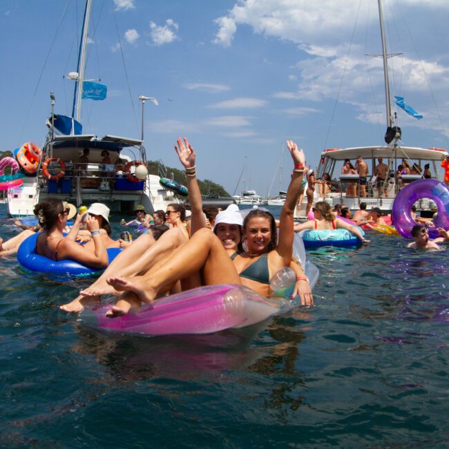 A lively group of people float on inflatable devices in a body of water, waving and enjoying their time. Several boats are anchored nearby under a partly cloudy sky. The scene is vibrant and everyone appears to be having fun at this inviting gathering, enjoying the beautiful day out on the water.