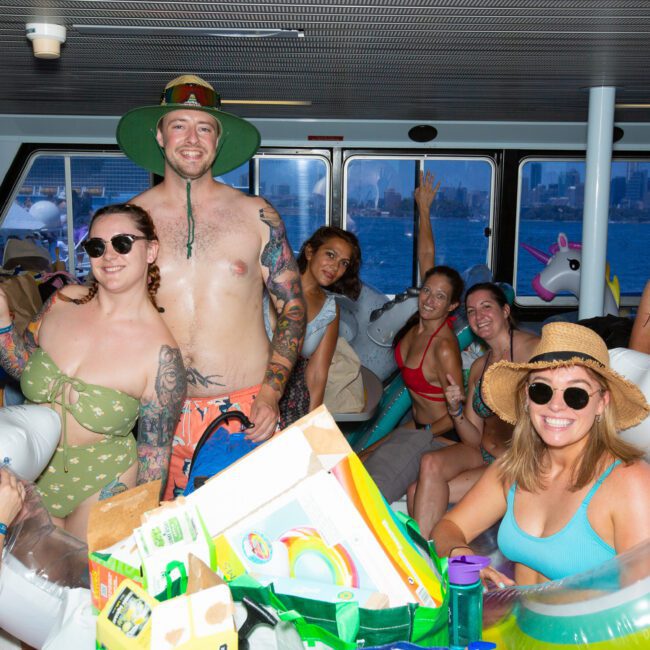 A group of people in swimwear are gathered on a boat, smiling and posing for the camera. Several are wearing hats and sunglasses, with inflatable pool toys around them. The background showcases large windows revealing an urban skyline over water, highlighting the vibrant cityscape.