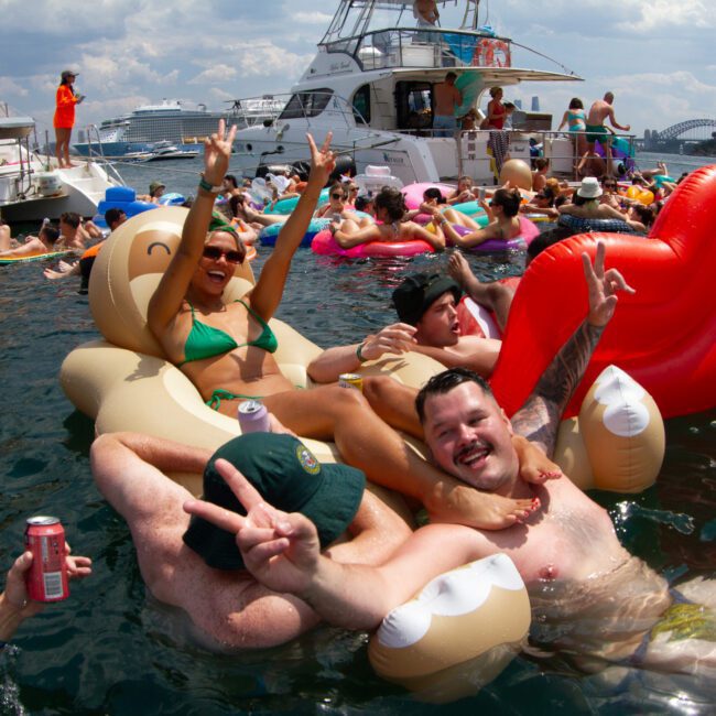 A lively group of people are enjoying a sunny day on the water, surrounded by inflatable floats and boats. Many are making peace signs and smiling at the camera. A woman in a green bikini sits prominently on a large unicorn-shaped float.