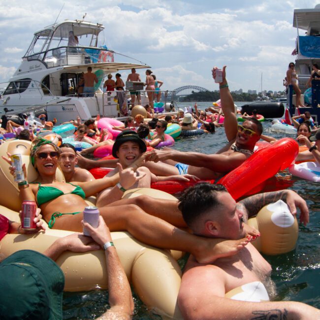 A large group of people enjoy a sunny day on the water, floating on colorful inflatables near yachts and boats. Some hold drinks, smile, and pose for the camera. The background features inflatable slides, a scenic bridge, and a blend of clouds and clear skies.