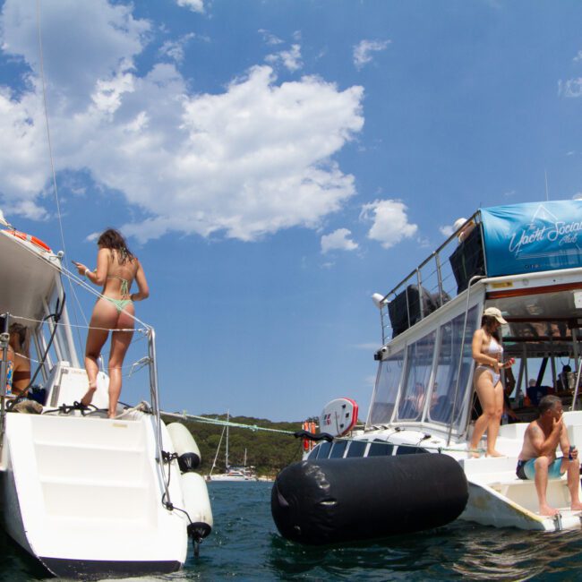 People are enjoying a sunny day on two docked boats in clear waters, some even snorkeling nearby. Some are climbing onboard while others are relaxing under an awning. The sky is clear with a few clouds, and lush green hills are visible in the background. The atmosphere is lively and fun.