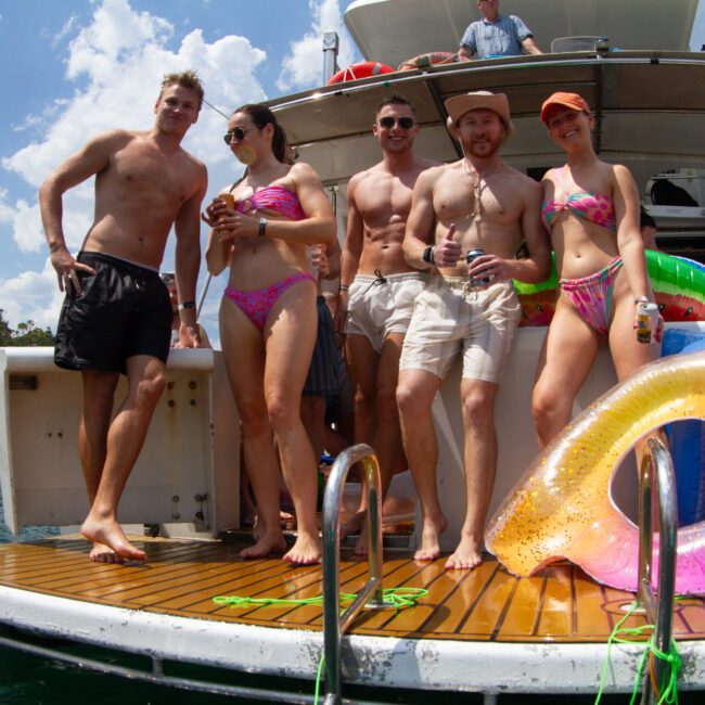 A group of five people in swimsuits stand on the deck of a boat, basking in the sunny weather. They are holding drinks and smiling. Vibrant inflatable rings are beside them, and jet skis zip across the water with other boats in the background.