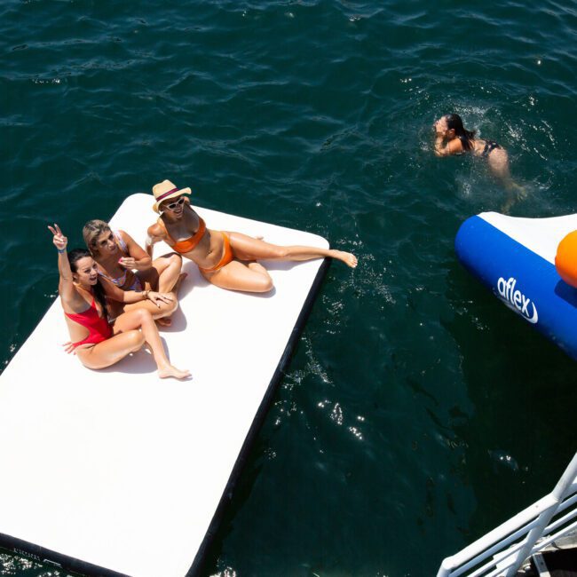 A group of three women in swimsuits are relaxing on a floating platform in the water. One woman is standing, another is lying down, and another is sitting with her arm raised. Nearby, a fourth person is swimming near an inflatable slide attached to a boat. The water glistens under the bright sun.