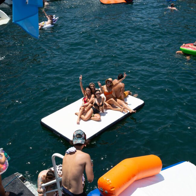 A group of people relax and pose for a photo on a floating mat in a body of water. Surrounding them, others are enjoying the water with inflatable tubes and floats. A boat is partially visible on the left side, adding to the lively and summery atmosphere of the scene.