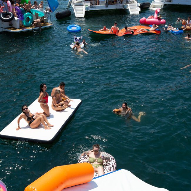 Several people enjoy a sunny day at a lake party with inflatable rafts and boats. Individuals are swimming, sunbathing, and lounging on vibrant pool floats shaped like a flamingo, watermelon, and doughnut. Boats are docked nearby, the lively atmosphere making it the perfect outdoor gathering for friends.