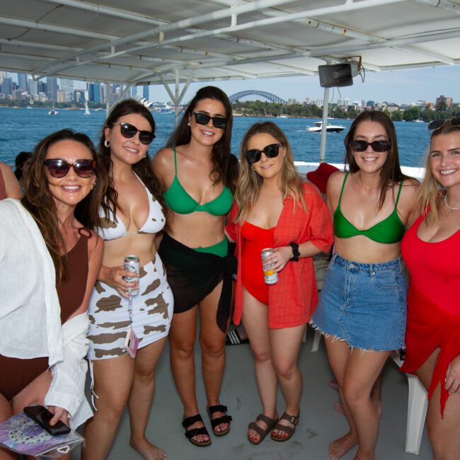 A group of seven women, dressed in various swimsuits and summer attire, stand together on a boat. They smile at the camera with a scenic water backdrop, showing a city skyline and bridge. Bright, sunny weather enhances the festive atmosphere as they prepare for their snorkeling adventure.
