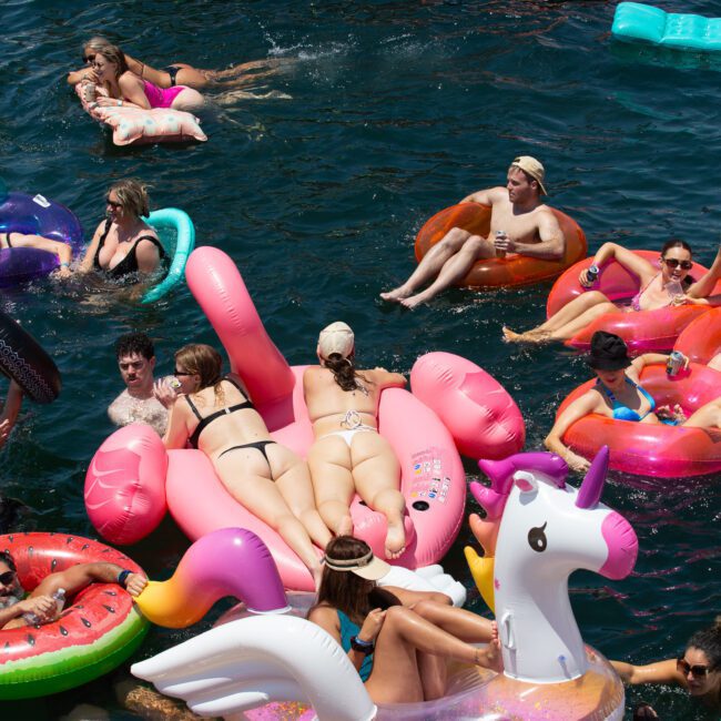 A lively scene of people enjoying a sunny day on the water. Individuals, both young and old, float on colorful inflatable pool floats, including unicorns, flamingos, and more. It's a crowded gathering at what appears to be a vibrant summer party on a picturesque lake or pool.