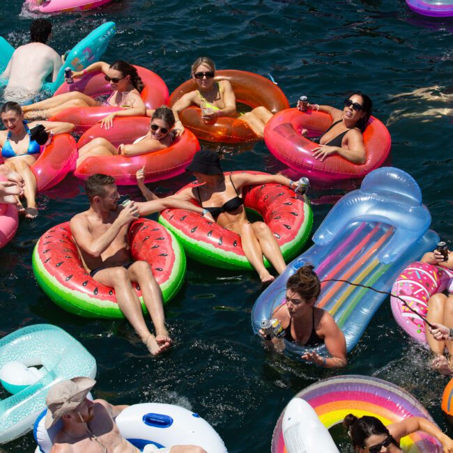 A group of friends are enjoying a sunny day floating on a serene lake. They use colorful inflatable rafts in various shapes, including a watermelon slice and a rainbow. Some socialize and hold drinks while others relax in the refreshing water.