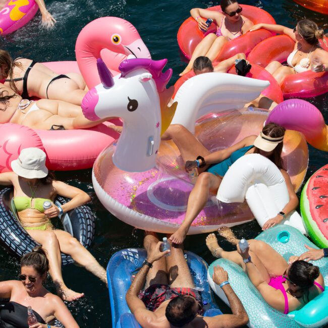 A crowded pool scene with numerous people relaxing on colorful inflatable floats, including a unicorn, a swan, and various fruit shapes. The sun is shining brightly, and the atmosphere appears festive and lively as everyone enjoys the day amidst cheerful splashes and laughter.