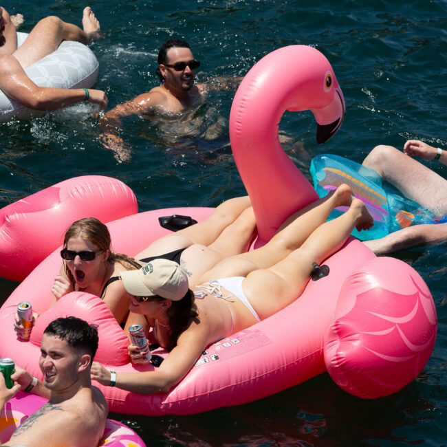 People enjoying a sunny day in the water, floating on inflatable rafts. Two women relax on a large pink flamingo float, holding drinks. Others float nearby on various other colorful inflatables, including a white inner tube and an inviting blue raft.