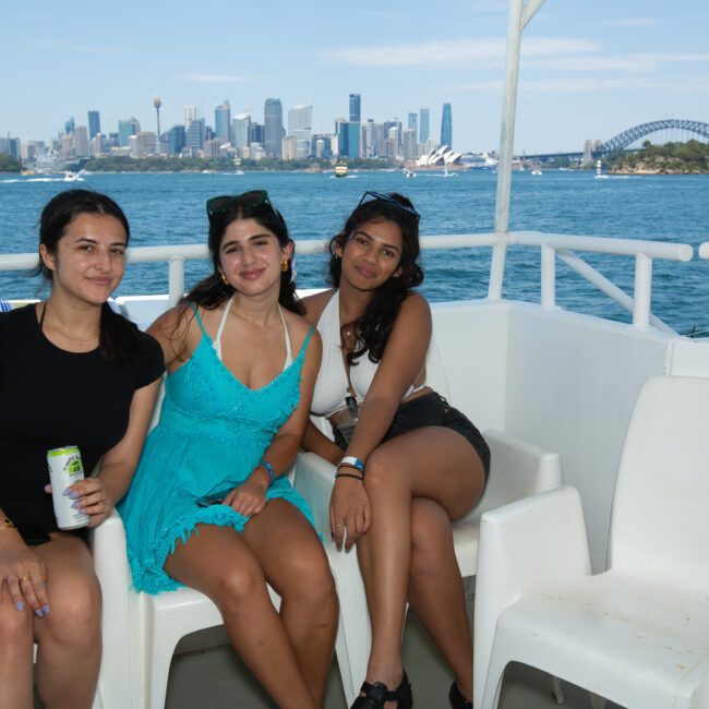 Three women are sitting on white chairs on the deck of a boat with a stunning cityscape, including the Sydney Opera House and Harbour Bridge, visible in the background. They are smiling and dressed in casual summer clothing. The sky is clear and blue.