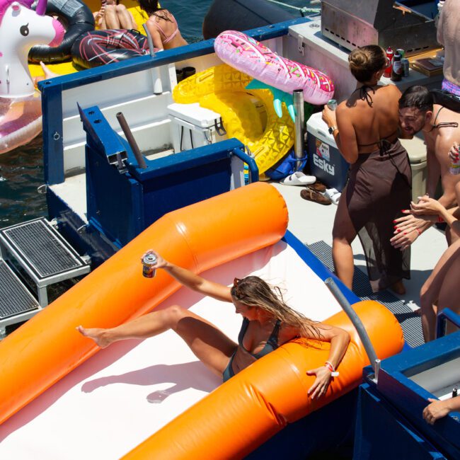 A group of people enjoy a sunny day on a boat, equipped with inflatable floats and a slide. One person slides down an orange inflatable slide into the sparkling water, while others relax and socialize around the boat. A barbecue grill adds to the festive atmosphere in the background.