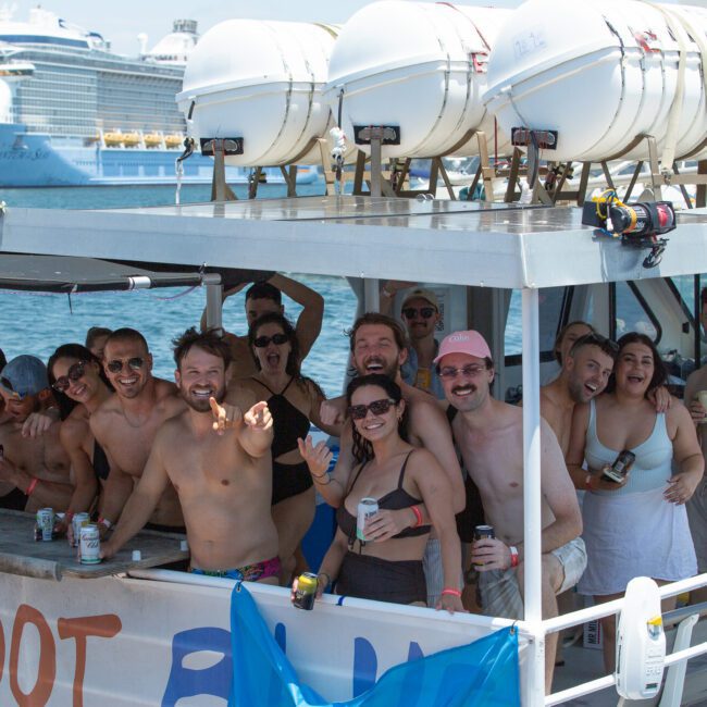 A group of people on a boat are smiling and holding drinks. They are dressed in swimwear and appear to be enjoying a sunny day on the water. A large cruise ship is visible in the background, adding to the excitement. The boat has a banner that partially reads "FOOT.