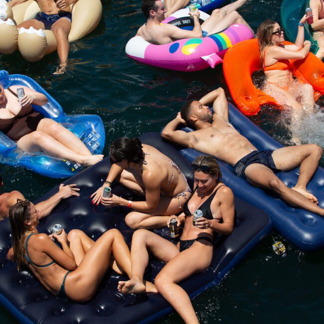 A group of people are enjoying a sunny day floating on a lake with a variety of inflatable loungers shaped like animals and air mattresses. They are relaxing, chatting, and holding drinks surrounded by calm water. Everyone in their colorful swimwear is soaking up the tranquil sun.