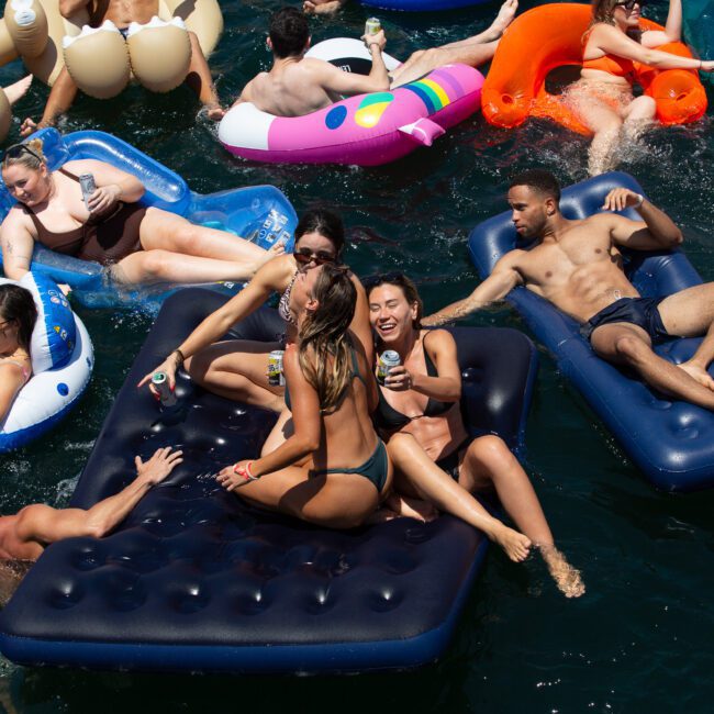People enjoying a sunny day on a lake, lounging on inflatable rafts and tubes. Some are chatting, laughing, and holding drinks as music plays in the background. The water is filled with various brightly colored floaties and the atmosphere is lively and fun.