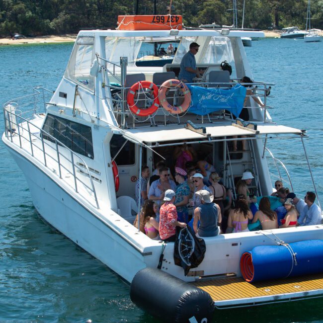 A group of people enjoying a sunny day on a white yacht anchored in a serene, blue-green water bay. The yacht has life rings, diving gear, and equipment on the deck. The backdrop includes a sandy shore and lush green trees. Some people are standing, and others are sitting.