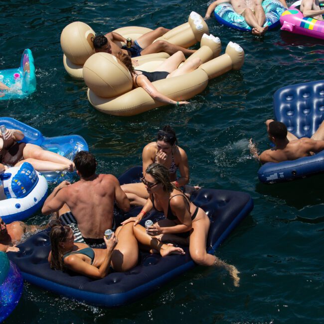 People relaxing on various inflatable floats in a body of water. Some individuals are lounging, while others are chatting. The scene is lively with a mix of brightly colored and creatively shaped floats, creating a joyful atmosphere. The deep blue water appears inviting and serene.