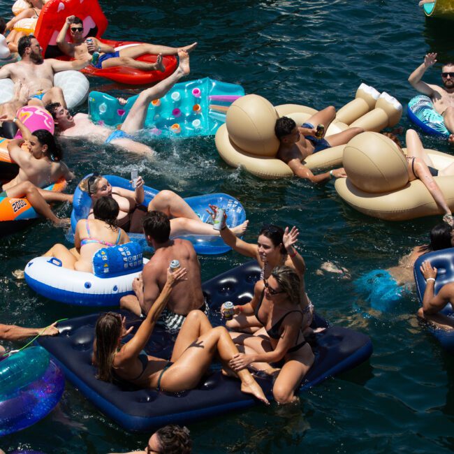 A lively crowd enjoys a sunny day on the water, floating on inflatable rafts of various shapes and colors. Some wave and smile at the camera, while others chat or lounge. The deep blue-green water enhances the festive atmosphere, making it a paradisiacal escape for all.