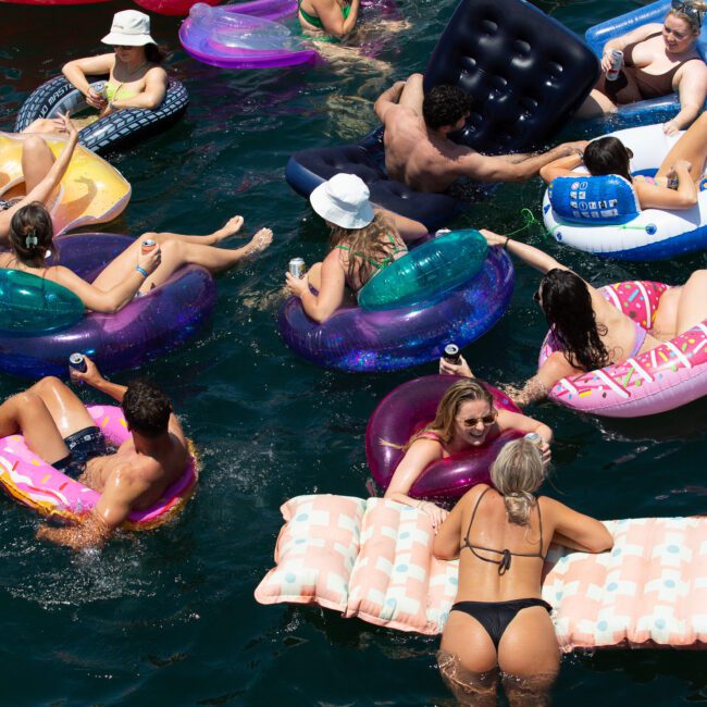 A group of people enjoying a sunny day on the water, lounging on various inflatable floaties including flamingos, donuts, and mattresses. The scene is vibrant with colorful floats, tropical swimwear, and a relaxed atmosphere.