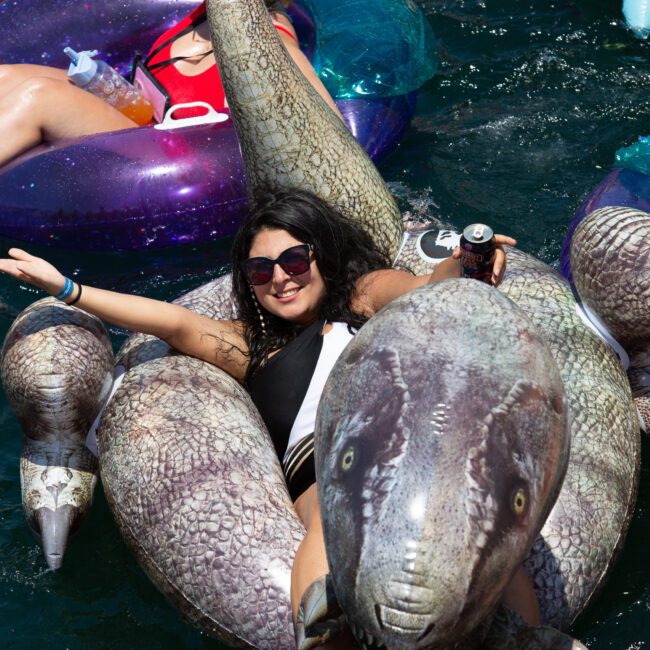 A woman lounges on a large inflatable pool toy shaped like a dinosaur, floating in water. She is smiling, wearing sunglasses, and holding a drink can. Other people on colorful inflatables are visible in the background, enjoying the sun-drenched scene.