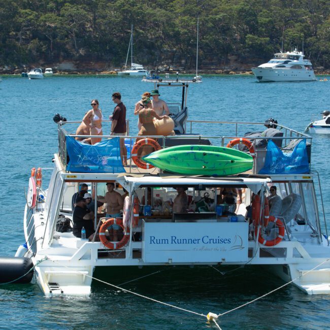 A boat named "Rum Runner Cruises" is positioned on a sparkling blue water body with people enjoying a sunny day on the deck. The boat carries an inflatable kayak on the roof and is surrounded by other boats near a scenic forested shoreline.