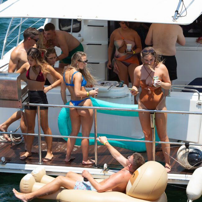 A group of people in swimwear socializes on the deck of a moored yacht on a sunny day. Some are holding drinks, while others stand at the railing. One person lounges on a float in the water next to the yacht. The scene is lively and relaxed.