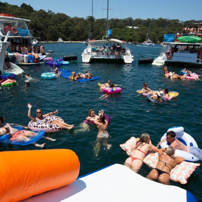 People are enjoying a sunny day on a lake, lounging on various inflatable floats and swimming. Multiple boats are anchored nearby, some with people socializing on the decks. The scene is lively, with lush greenery visible in the background and paddleboarders drifting peacefully.