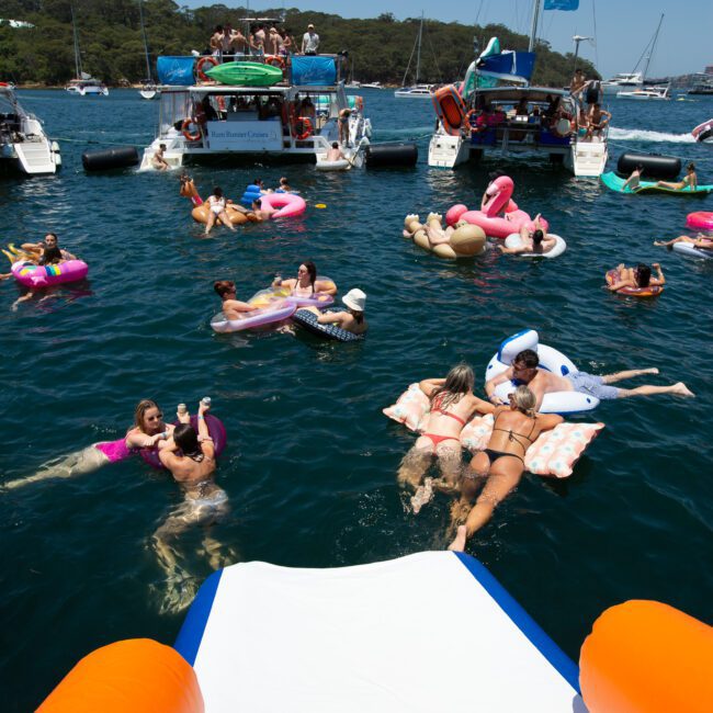 People floating on colorful inflatable tubes and toys in a body of water, surrounded by anchored boats and yachts. The scene is lively with a mixture of relaxation and play. A slide extends from a boat into the water, bathed in clear blue skies, with scenic greenery providing a picturesque backdrop.