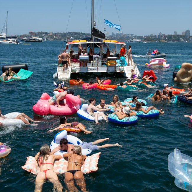 A group of people is enjoying a sunny day on the water, floating on various inflatable pool toys near a yacht. The calm, blue water reflects the clear sky, with more boats and a stunning cityscape in the background. Everyone appears relaxed and having fun.