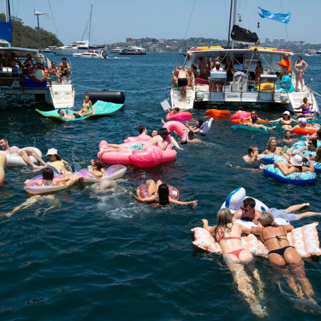 A lively scene on the water features numerous people enjoying a sunny day on inflatable devices. Several boats are anchored nearby, with folks socializing and swimming. The image radiates a fun, festive atmosphere with clear blue water, an inviting coastline, and a clear sky in the background.